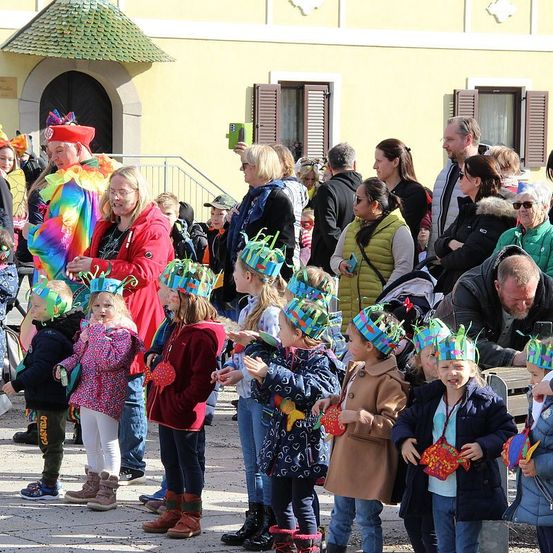 Eine Menschenmenge versammelt sich im Freien, mehrere Kinder tragen festliche Hüte und halten fischförmige Objekte. Ein Mann in bunter Kleidung steht in ihrer Nähe. Hinter der Menge ist ein Gebäude mit Fensterläden und einer Treppe zu sehen.