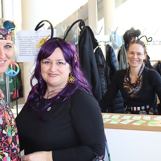 Drei Frauen in bunten Outfits, eine mit lila Haaren und Biene-Kopfbedeckung, stehen vor einem Tresen. Dahinter hängen Kleidung an Haken und ein Zettel ist an der Scheibe befestigt.