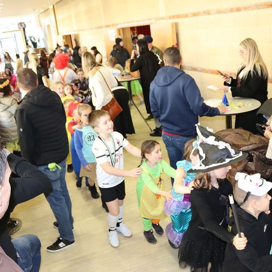 Innenhalle mit Kindern in Kostümen, Erwachsenen und einem Tisch mit Gegenständen. Einige tragen Hüte und Masken. Ein Junge in einem weißen Shirt hält die Hand eines Mädchens.