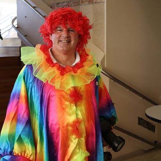 Ein Mann in einem farbenfrohen Clownskostüm mit einem Rüschenkragen steht neben einer Treppe und hält eine Kamera.