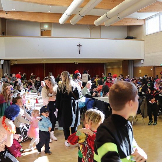 Ein großer Saal mit Holzboden, Menschen in Kostümen, Tische mit Essen, eine lächelnde Frau, spielende Kinder und ein Kreuz an der Wand.