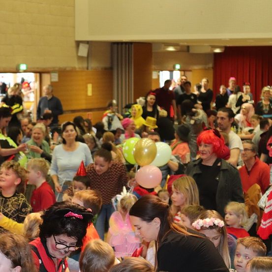 Eine Menschenmenge, vermutlich auf einem Karneval, besteht aus Erwachsenen und Kindern in verschiedenen Kostümen und Hüten. Einige halten Luftballons, andere tragen Brillen und Masken. Die Umgebung ist drinnen mit einem roten Vorhang.
