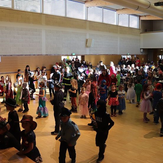 Eine große Gruppe von Kindern in Kostümen, darunter Superhelden und Feuerwehrleute, tanzt in einem Saal mit Holzböden. Einige Erwachsene sind ebenfalls anwesend, und der Saal hat weiße Wände und Deckenleuchten.