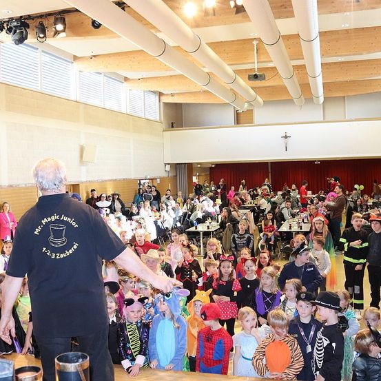 Ein Mann steht auf einer Bühne mit Kindern in verschiedenen Kostümen. Er scheint für sie aufzutreten. Die Kinder hören aufmerksam zu. Der Ort hat Holzbalken an der Decke und einen hängenden Projektor. Der Boden ist aus Holz und die Wände sind weiß. Es gibt viele Menschen, die auf Stühlen sitzen. Einige von ihnen essen.