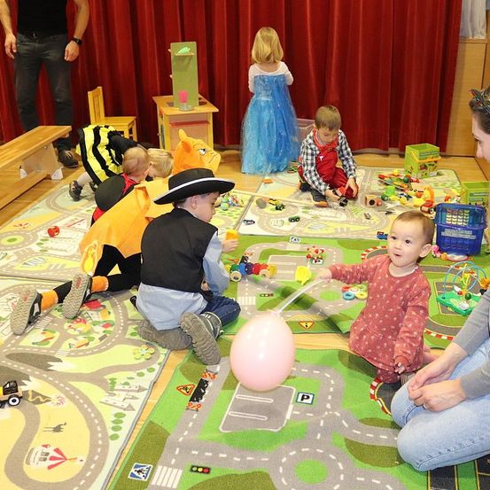 Eine Gruppe von Kindern spielt mit Spielzeug auf einem straßenthematischen Teppich. Ein Baby in einem rosa gepunkteten Kleid spielt mit einem rosa Ballon. In der Nähe spielen ein Mädchen in einem blauen Kleid und ein Junge in einem karierten Hemd mit Spielzeug. Ein weiteres Kind ist als Biene verkleidet.