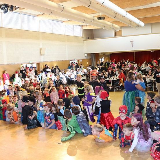 Eine große Menschenmenge aus Kindern und Erwachsenen in verschiedenen Kostümen versammelt sich drinnen, möglicherweise für eine Kostümparty. Viele sitzen, während andere stehen und beobachten. Tische sind mit Essen und Getränken gedeckt.