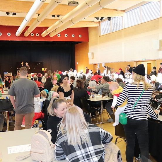 Ein Indoor-Event zeigt eine Menschenmenge, einige sitzen und andere stehen und unterhalten sich. Tische sind mit verschiedenen Gegenständen beladen und eine Frau hält ein Baby in einem Tragetuch. Der Hintergrund umfasst eine Bühne mit einem Lautsprecher, und die Decke hat mehrere Rohre.