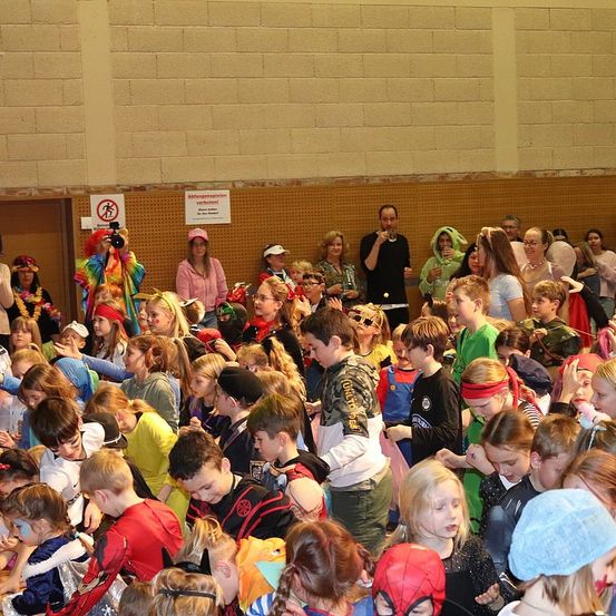 Ein überfüllter Raum mit Kindern in verschiedenen Kostümen, einige mit Gesichtsbemalung, ein Mann mit einer Kamera und Erwachsene, die sich im Hintergrund bewegen.
