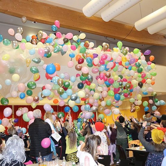 Ein Raum voller Menschen bei einer Veranstaltung. Viele Luftballons in verschiedenen Farben schweben in der Luft. Einige Leute heben ihre Hände. Ein paar Leute halten Ballons in den Händen.