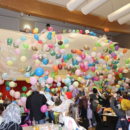 Eine Menschenmenge, einschließlich Kinder, versammelt sich in einem Raum voller bunter Luftballons, die von der Decke hängen. Einige halten Luftballons, während andere danach greifen. Ein Mann in einem Hut steht auf einer Plattform.