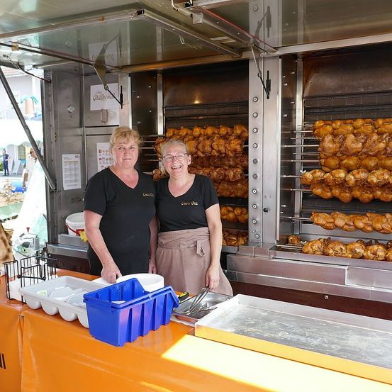 Zwei Frauen stehen lächelnd hinter einem Imbiss-Counter und haben Geschirr und Utensilien auf dem Tisch. Dahinter befinden sich Öfen mit gebratenem Hähnchen.