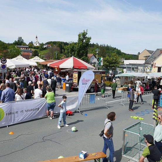 Ein Straßenmarkt mit Zelten, Bannern und einem Fußballspiel. Menschen gehen herum, und es gibt Straßenlaternen, Bäume und eine Kirche in der Ferne.