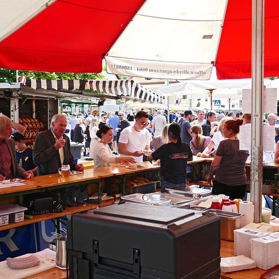 Ein geschäftiger Imbissstand mit einem rot-weiß gestreiften Baldachin. Mehrere Menschen stehen und sitzen um Tische herum, essen und trinken. Es gibt Tabletts, Flaschen und andere Gegenstände auf den Tischen.