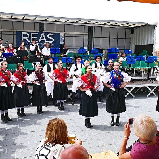 Eine Gruppe von Künstlern in traditioneller Kleidung steht auf der Bühne, während eine Band hinter ihnen spielt. Das Publikum beobachtet sie und einige machen Fotos. Die Bühne hat einen Hintergrund mit dem Wort 'REAS'.