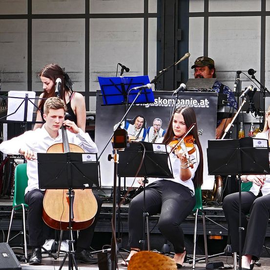 Eine Gruppe von Musikern tritt auf einer Bühne mit Hintergrund auf. Ein Cellist, Geiger und andere spielen. Ein Schild mit Text steht dahinter.