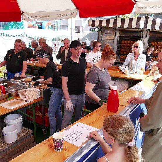 Eine Gruppe von Menschen in schwarzen und grauen Shirts bereitet Essen an einem Essensstand vor. Ein Mann und eine Frau stehen vor der Theke, und ein Kind sitzt vor ihnen. Es gibt Flaschen, eine Schüssel und ein Tablett mit Essen auf der Theke. Es gibt Regenschirme, und andere Essensstände sind in der Gegend.