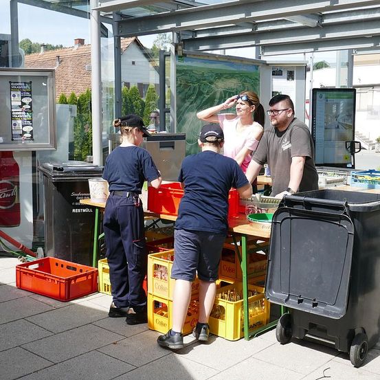 Vier Menschen stehen in einem Buswartehäuschen, drei in Uniformen. Einer hält einen Mülleimer, zwei andere schauen sich Gegenstände auf einem Tisch an. Eine Frau in einem rosa Hemd hält eine Karte.
