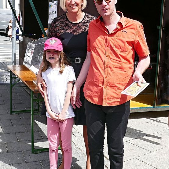Eine Frau und ein Mann stehen lächelnd mit einem Mädchen vor einem Holztisch. Das Mädchen trägt einen rosa Hut und rosa Hosen. Hinter ihnen ist eine offene Tür eines Standes.