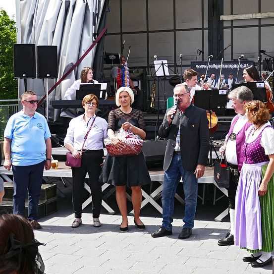Eine Gruppe von Menschen steht auf einer Bühne mit einer Band. Ein Mann hält ein Mikrofon, während eine Frau einen Korb hält. Hinter ihnen spielt ein Pianist, und andere sind mit Musikinstrumenten auf der Bühne.
