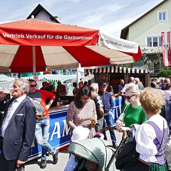 Ein geschäftiger Outdoor-Markt mit mehreren Ständen und Menschen. Ein Regenschirm mit der Aufschrift 'Vertrieb Verkauf für die Gastronomie' ist vorhanden. Einige Leute halten Getränke in der Hand und andere stöbern durch die Stände. Ein Kinderwagen wird von einer Frau geschoben.