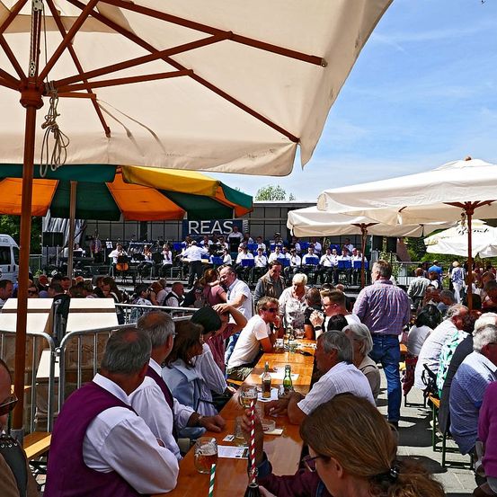 Eine große Gruppe von Menschen versammelt sich unter Schirmen um Tische, einige tragen Hüte und Brillen. Eine Band spielt auf einer Bühne, mit Leuten in Stühlen und einer Metallbarriere in der Nähe. Der Himmel ist klar mit einigen Wolken.