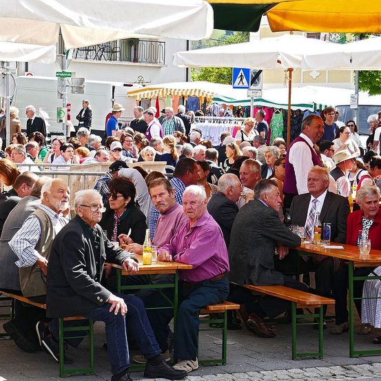Eine Gruppe älterer Menschen sitzt auf Bänken bei einer Veranstaltung im Freien. Sie sind unter Schirmen und einige tragen Brillen. Einige unterhalten sich und andere schauen etwas an.