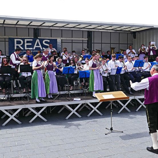 Ein Orchester spielt auf einer Bühne, Musiker spielen verschiedene Instrumente. Der Dirigent steht vorne, und ein Banner mit Text ist sichtbar.