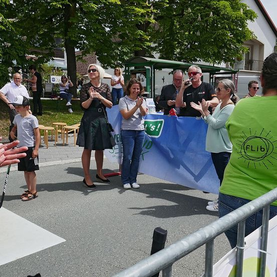 Eine Gruppe von Menschen versammelt sich im Freien, einige stehend und klatschen, andere sitzend oder gehend. Ein Banner mit dem Text 'Es ist!' wird von zwei Frauen gehalten. Im Hintergrund befindet sich ein Gebäude und ein Baum.