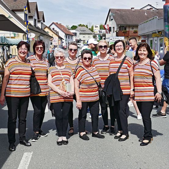 Eine Gruppe von Frauen in bunt gestreiften T-Shirts steht auf einer Straße und lächelt für ein Foto. Sie tragen schwarze Hosen und Lederschuhe. Hinter ihnen befinden sich Gebäude und Bäume.
