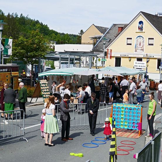 Eine Straßenszene mit verschiedenen Menschen und einem lebhaften Outdoor-Markt. Einige Personen spielen Spiele, während andere Stände erkunden. Ein gelber Imbisswagen und Banner sind sichtbar, mit einem Gebäude im Hintergrund.