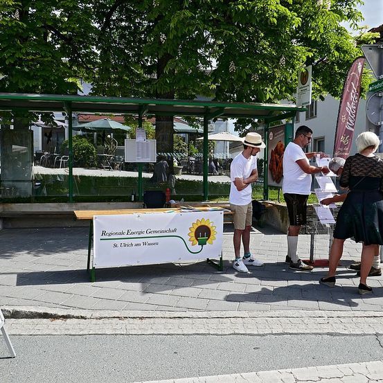 Zwei Männer und zwei Frauen stehen neben einem Tisch mit einem Banner, auf dem steht Regionale Energie Gemeinschaft, St. Ulrich am Waasen. Sie sind vor einer Bushaltestelle.