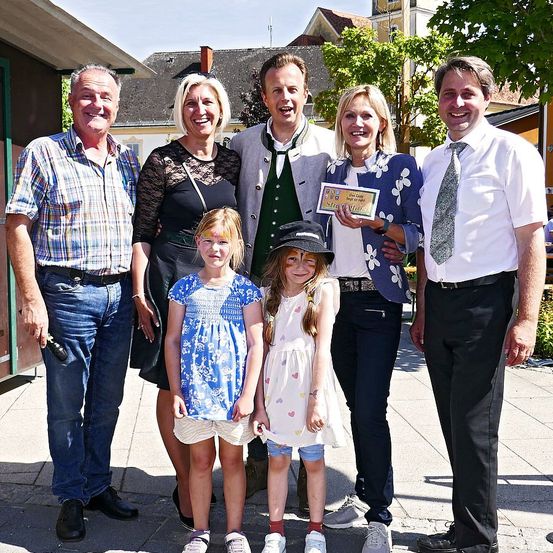 Eine Gruppe von Menschen, einschließlich Erwachsener und Kindern, stehen zusammen und lächeln für ein Foto. Der Mann in der Mitte hält ein Zertifikat. Hinter ihnen stehen Bäume und Gebäude.