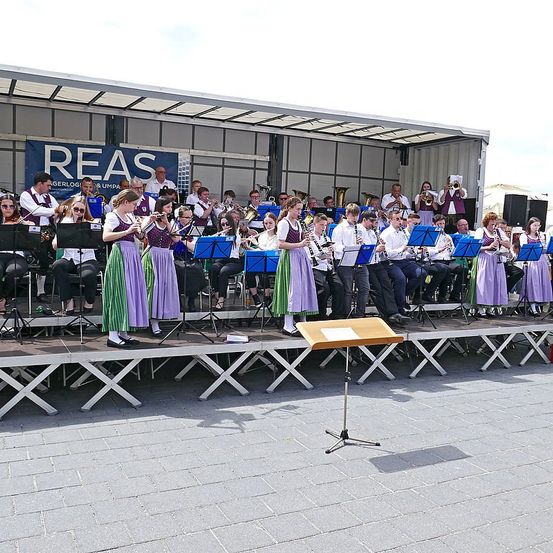 Ein Orchester spielt auf einer Bühne mit Musikinstrumenten. Die Bühne hat einen Holzboden und einen Metallrahmen. Die Bandmitglieder sind in lila und grünen Kostümen gekleidet. Einige sitzen, andere stehen. Dahinter befindet sich ein Banner mit dem Text 'REAS'.