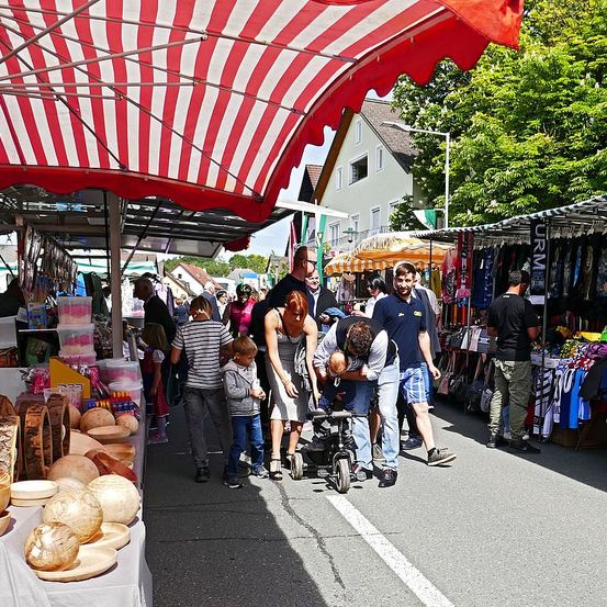 Ein Markt mit einem rot-weiß gestreiften Vordach bietet verschiedene Stände. Menschen schlendern mit Kindern, einige schieben Kinderwagen. Ein Mann beugt sich über einen Kinderwagen. In der Nähe gibt es Tische mit Holzschüsseln.