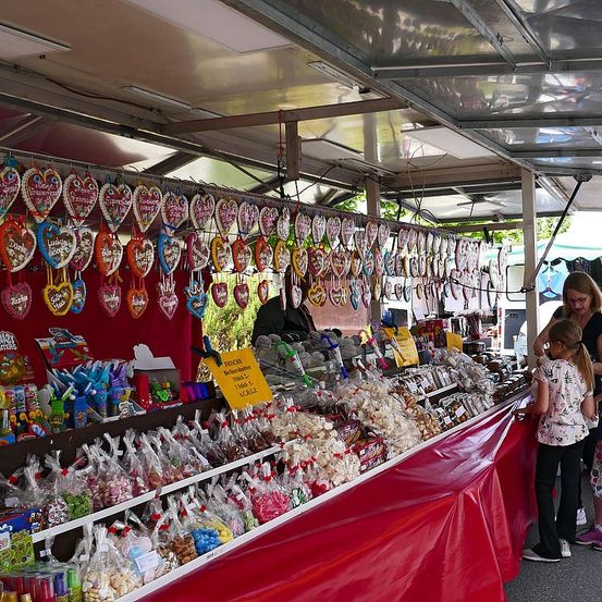 Ein überdachter Marktstand ist gefüllt mit bunten herzförmigen Keksen und verschiedenen Süßigkeiten. Mehrere herzförmige Ornamente hängen von der Decke. Eine Frau und ein Mädchen stehen vor dem Stand.