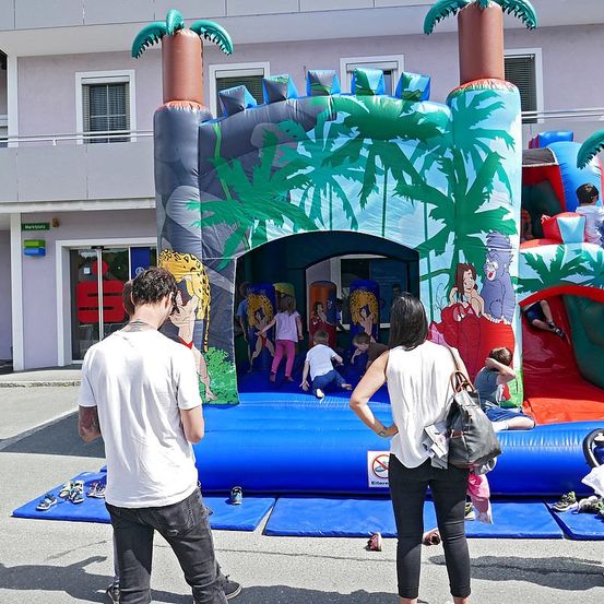 Eine Gruppe von Kindern spielt in einem großen aufblasbaren Schloss mit Palmendesigns. Ein Mann und eine Frau stehen in der Nähe. Einige Schuhe sind auf dem Boden platziert. Ein Gebäude mit Glastüren ist im Hintergrund.
