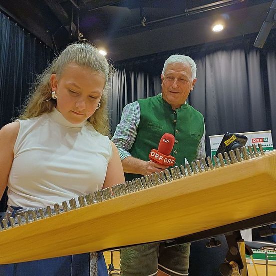 Eine Frau spielt eine Zither, während ein älterer Mann ein Mikrofon vor ihr hält. Die Zither wird von der Frau mit beiden Händen gehalten.