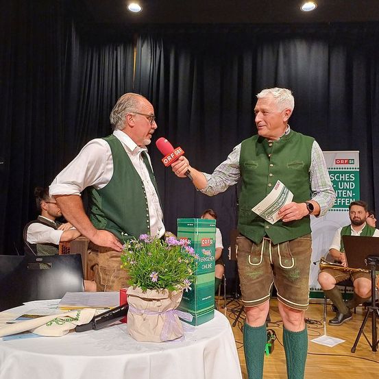 Zwei Männer werden auf der Bühne mit einem Mikrofon interviewt, halten ein Papier und ein Buch. Ein Tisch mit einem Blumenvase steht vor ihnen. Dahinter befindet sich ein schwarzer Vorhang und ein Mann sitzt auf einem Stuhl.