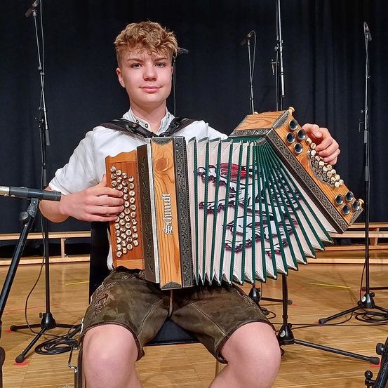 Ein junger Mann sitzt auf einem Stuhl und spielt ein antikes Akkordeon mit aufwendigen Verzierungen. Er trägt ein weißes Hemd und braune Shorts, vor ihm stehen Mikrofone. Der Ort scheint eine Bühne mit schwarzem Hintergrund zu sein.