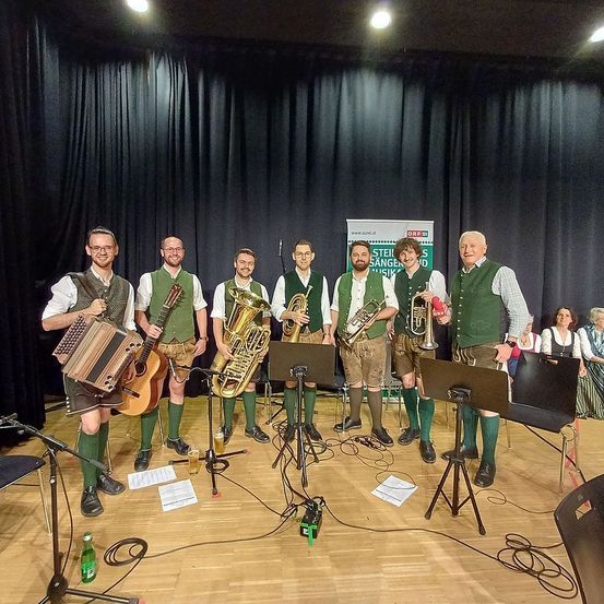 Eine Gruppe von Musikern steht mit Musikinstrumenten auf einer Bühne, ein Banner hinter ihnen trägt den Schriftzug Steirer Sangerbund Musik.