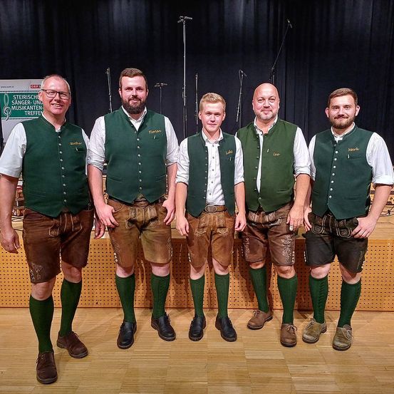 Fünf Männer in traditioneller Kleidung posieren für ein Foto auf einer Bühne, mit Mikrofonen und einem Banner im Hintergrund.