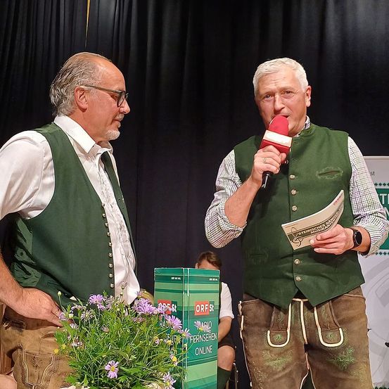 Zwei Männer in traditioneller Kleidung stehen auf der Bühne, einer hält ein Mikrofon und der andere schaut ihn an. Ein Blumenstrauß steht vor ihnen.