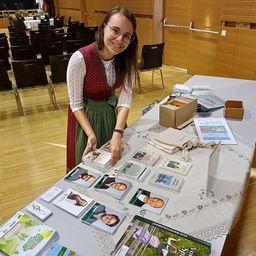 Eine Frau steht in einem Konferenzsaal an einem Tisch, trägt eine Brille und ein traditionelles Kleid. Der Tisch ist mit einer Spitzentischdecke bedeckt und hält Bücher, Broschüren und eine Tasche. Der Hintergrund zeigt Reihen von Stühlen und eine Holzwand.
