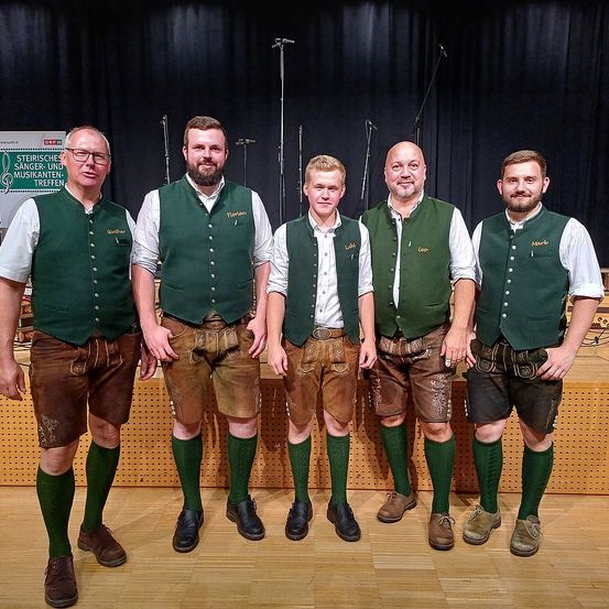 Fünf Männer, gekleidet in traditioneller deutscher Kleidung, einschließlich grüner Westen und brauner Shorts, stehen auf einem Holzboden. Sie lächeln und scheinen für ein Foto zu posieren. Hinter ihnen befindet sich ein schwarzer Vorhang mit einem Schild, das 'Steirische Sänger- und Musikantentreffen' besagt.