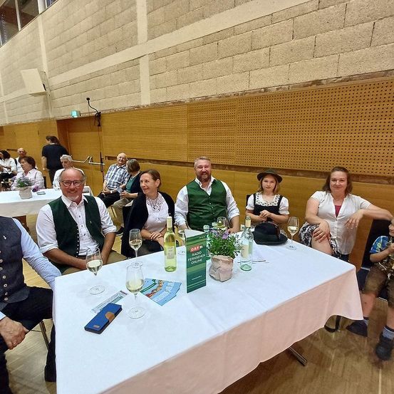 Eine Gruppe von Menschen in traditioneller Kleidung sitzt an einem Tisch, umgeben von Weinglasern und einer Blumenvase. Sie befinden sich in einem Raum mit Holzwänden und -böden.