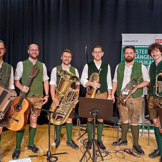 Sechs Männer in grünen Westen und Shorts posieren für ein Foto mit Musikinstrumenten. Sie haben Tuba, Trompete und Akkordeon. Sie stehen vor einem Notenständer und einem schwarzen Hintergrund.