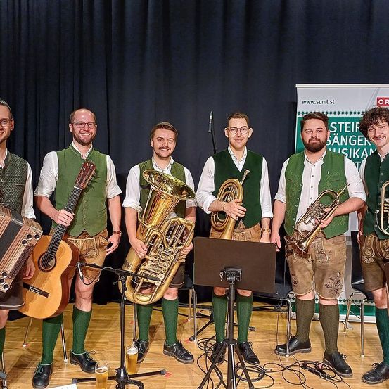 Fünf Männer in traditioneller deutscher Kleidung stehen auf einer Bühne und halten Musikinstrumente, darunter Gitarren, Tubas und Trompeten. Sie sind von Stühlen und einem Notenständer umgeben, und der Hintergrund zeigt einen schwarzen Vorhang.