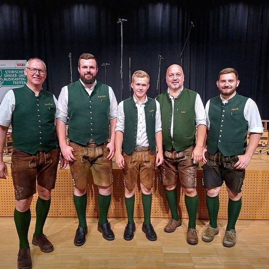 Fünf Männer in traditioneller deutscher Kleidung stehen auf der Bühne und lächeln für die Kamera. Sie tragen grüne Westen und braune Hosen, zwei von ihnen mit Brille. Die Bühne ist mit schwarzen Vorhängen und Mikrofonen geschmückt.