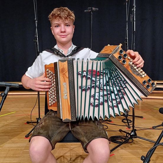 Ein junger Mann sitzt auf einem Stuhl und hält ein Akkordeon, möglicherweise für ein Foto. Er trägt ein weißes Hemd und grüne Hosenträger. Ein Mikrofon auf einem Ständer steht hinter ihm.