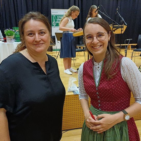 Zwei Frauen stehen zusammen, eine in einem schwarzen Shirt, die andere in einem traditionellen Kleid, und lächeln. Hinter ihnen spielt eine Gruppe von Musikern, mit einem Banner, das 'Steirische Gams' im Hintergrund zeigt.